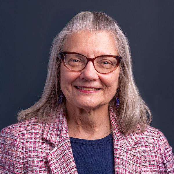 Portrait of a smiling older woman with gray hair, red glasses, wearing a pink plaid blazer over a navy top, against a dark background.
