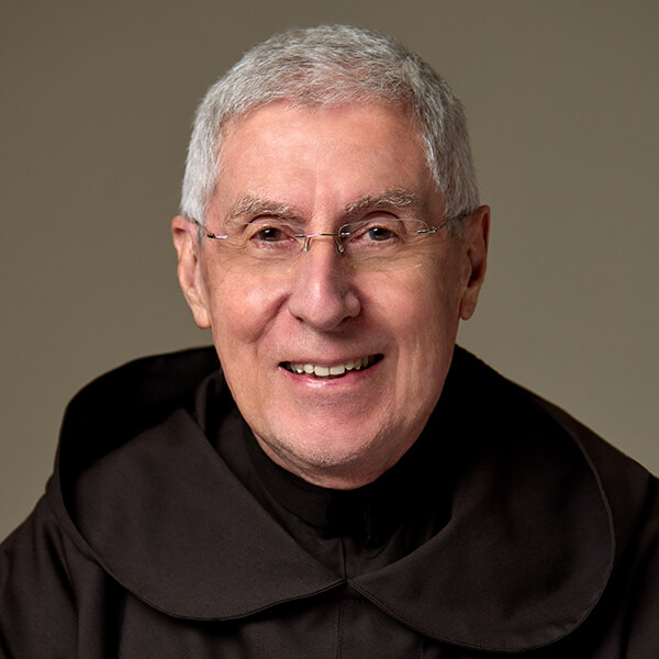 Smiling elderly man with short gray hair and glasses wearing a black clergy/academic robe, against a neutral background.