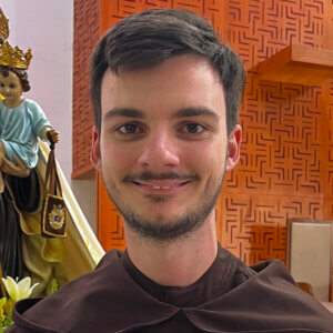 Smiling young man in brown religious habit indoors, with a wooden patterned wall and a statue in the background