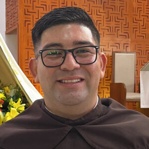 Portrait of a man with black hair and glasses, wearing a brown clerical robe, smiling in an indoor setting with orange patterned wall in the background.