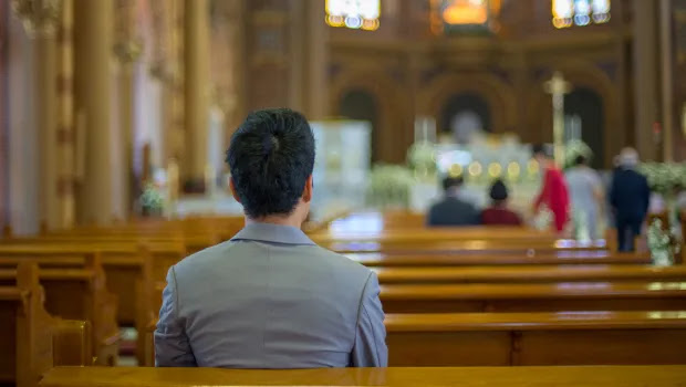 Back view of a man in a gray suit seated in a wooden church pew, facing the blurred altar at the front.