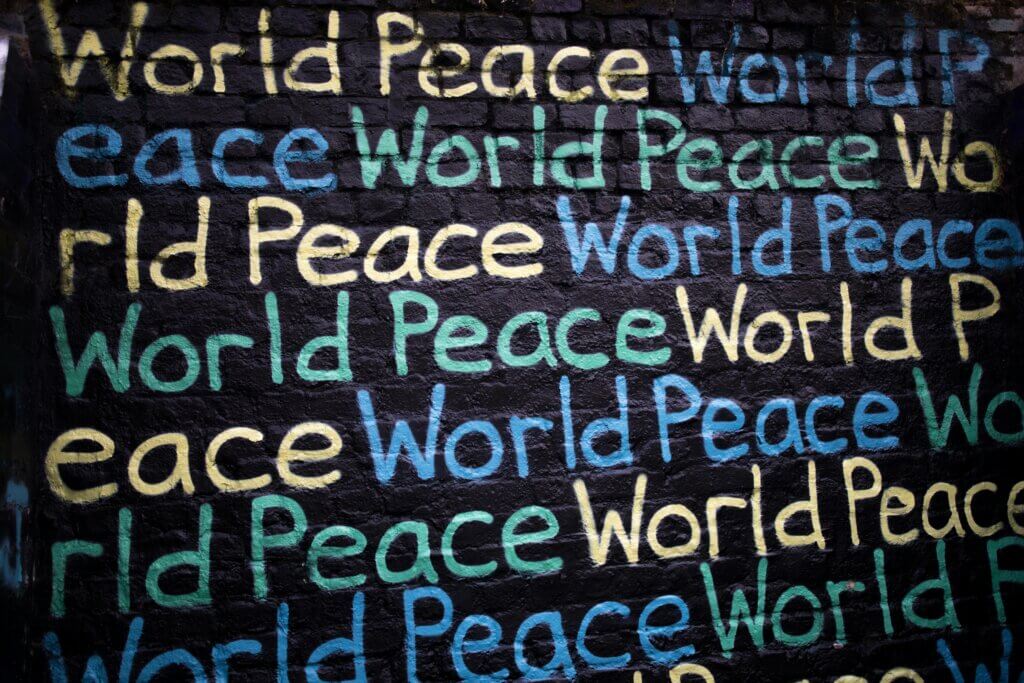 Colorful graffiti wall with the word 'peace' repeated in green, yellow, blue, and white on a dark brick surface.