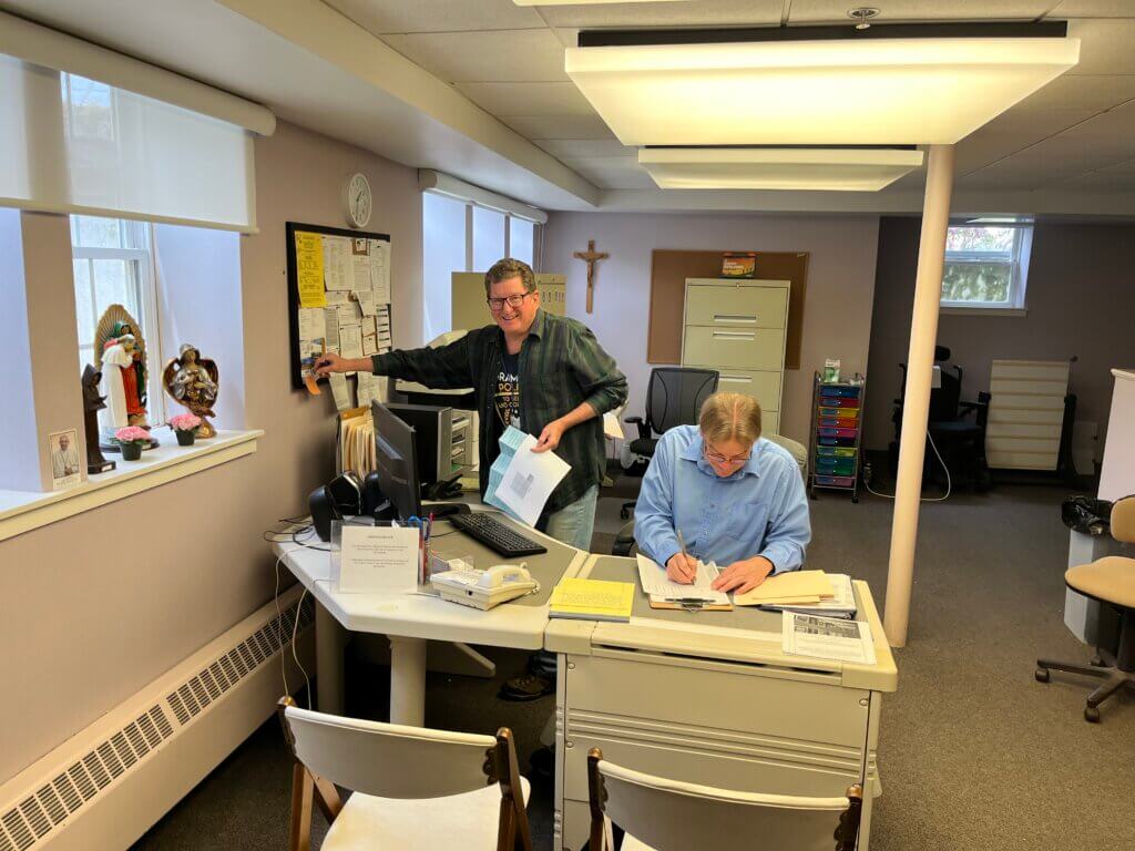 Man in a green plaid shirt hands papers to a seated colleague at a reception desk in a small office.