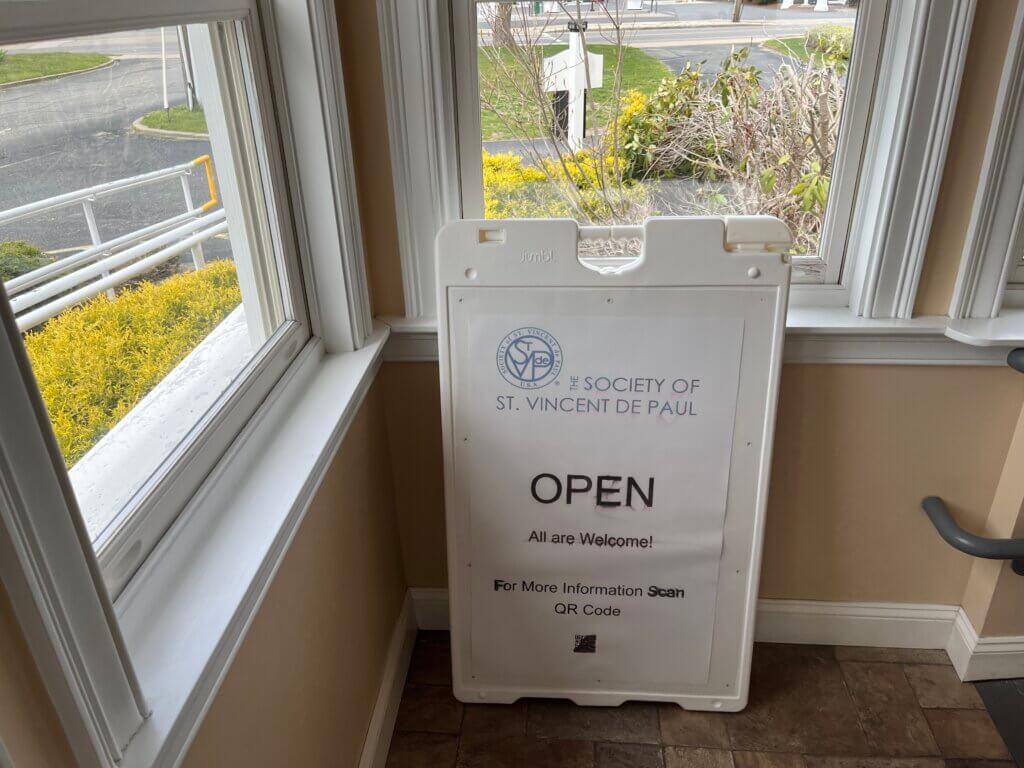 Indoor view of a white donation sign holder with 'OPEN' text, standing by a window; outside shows yellow bushes and a parking lot.
