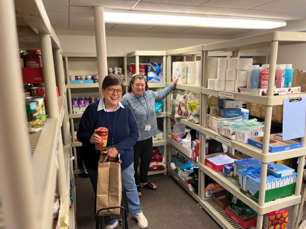 Two smiling women in a cluttered stockroom aisle, one holding a jar and a paper bag on a cart amid shelves of household supplies.