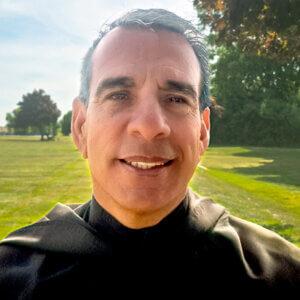 Close-up selfie of a smiling man in black clerical attire outdoors on a sunny day, with a green park behind him.