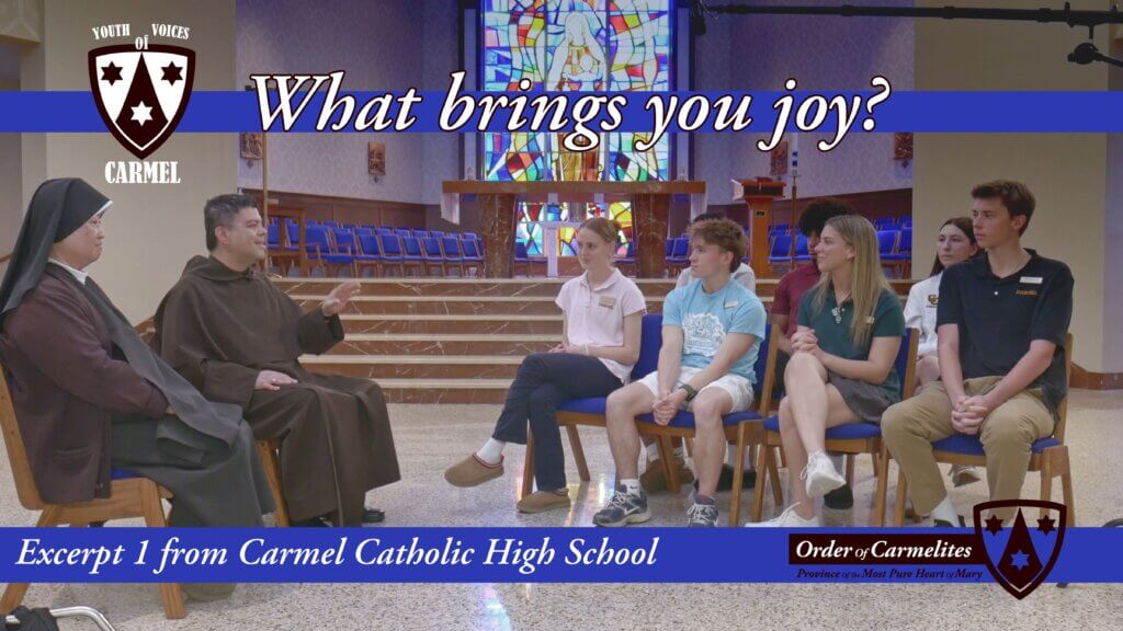 Group of high-school students seated in a church chapel, listening to two friars, with a blue banner reading 'What brings you joy?' and a Carmel crest logo on the left.