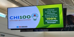 Digital information screen at a transit station displaying CHI100 logo and a message about 100% renewable energy.