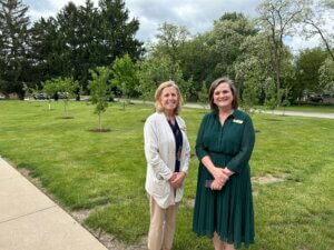 Two women standing smiling on a park lawn with trees in the background wear business‑casual outfits.