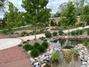 Campus courtyard with a stone statue fountain surrounded by trees and brick buildings; people walk along paved paths by a koi-free pond with rocks and plants nearby.