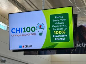 Digital sign promoting Chi100 at Midway Airport: 'Please Enjoy Your Midway Experience' with 100% Renewable Energy message and Chicago branding.