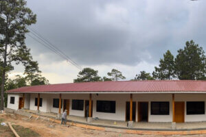 A long single-story building with a red metal roof, white walls, and wooden columns, under construction with workers on a dirt site and trees in the background under a cloudy sky.