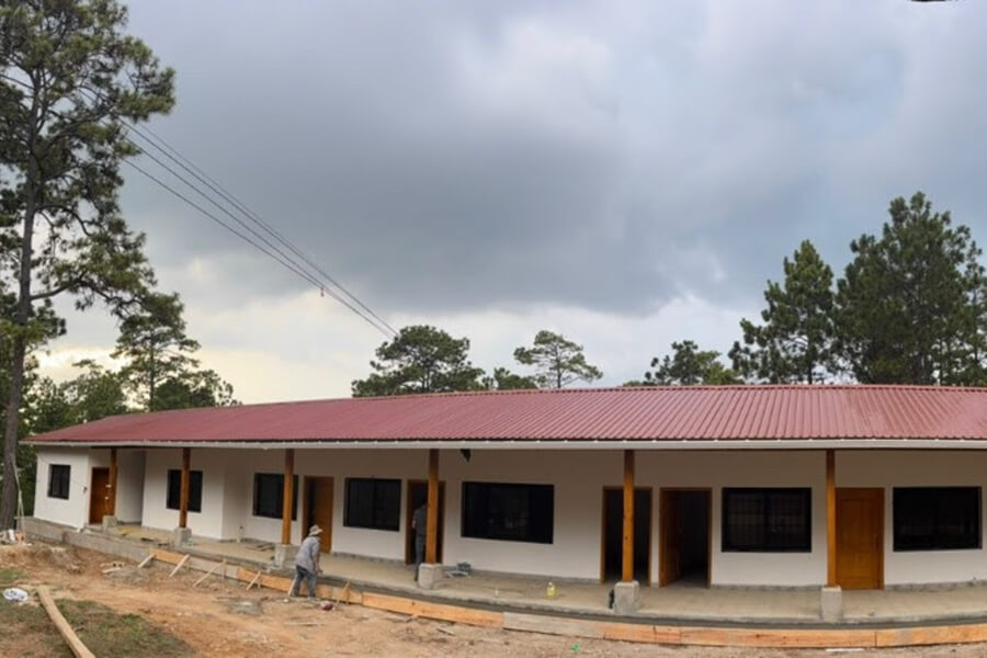A long single-story building with a red metal roof, white walls, and wooden columns, under construction with workers on a dirt site and trees in the background under a cloudy sky.