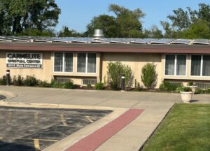 Front view of the Carmelite Spiritual Center building with a sign that reads 'Carmelite Spiritual Center, 8419 Main Entrance', sidewalk, and landscaped plants.