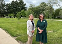 Two women standing on a grassy park with trees, smiling at the camera.