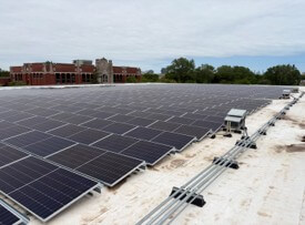 Rows of solar panels installed on a flat white rooftop with a building and trees in the background.