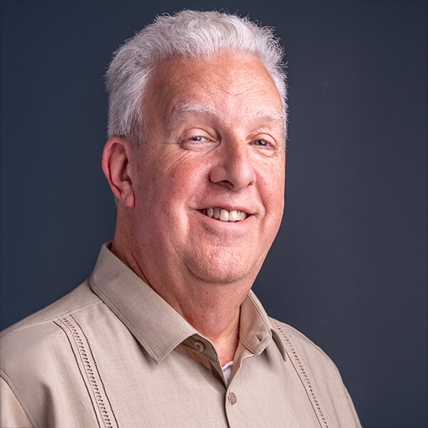 Smiling older man with gray hair wearing a beige collared shirt against a dark background; portrait-style headshot.
