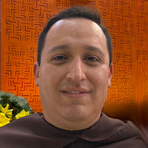 Close-up of a man in a brown robe smiling, with an orange patterned wall behind him.