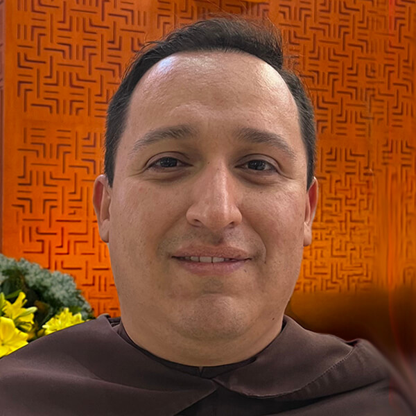 Close-up of a man in a brown robe smiling, with an orange patterned wall behind him.