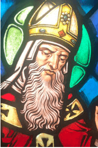 Stained-glass window depicting a bearded bishop wearing a gold mitre and red robes, surrounded by colorful panes.